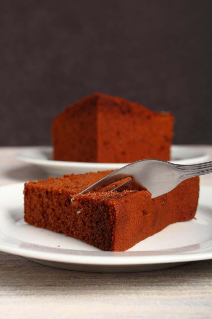 A slice of gingerbread cake on a plate with a cup of tea in the background.