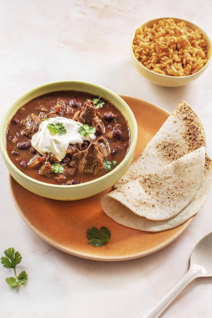 A well-rounded meal with slow cooker chili con carne, Spanish rice, and whole wheat tortillas on an earthy-toned plate.