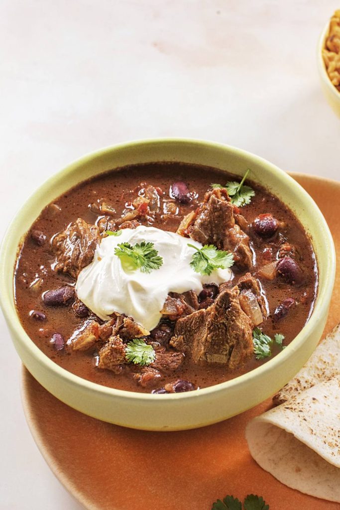 Hearty slow cooker chili con carne topped with a dollop of sour cream and fresh cilantro in a green bowl.