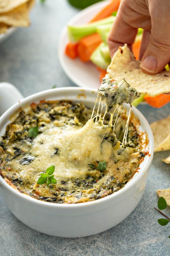 Hand lifting a chip with spinach and artichoke dip, showing melted cheese.