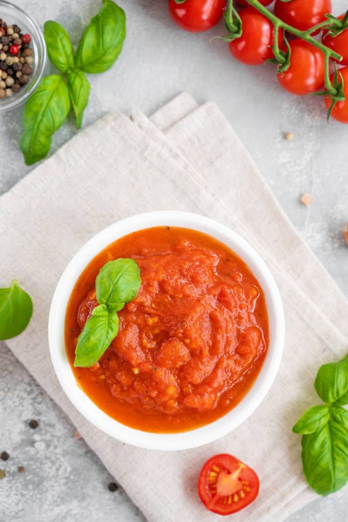 A bowl of red marinara sauce next to fresh basil.