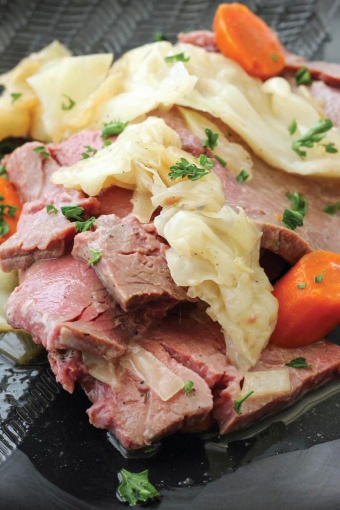 Slices of juicy corned beef with cabbage and carrots on a dark plate, garnished with parsley.