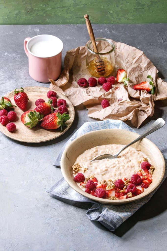 A bowl of creamy porridge topped with raspberries and nuts next to fresh fruit and honey on a rustic table.