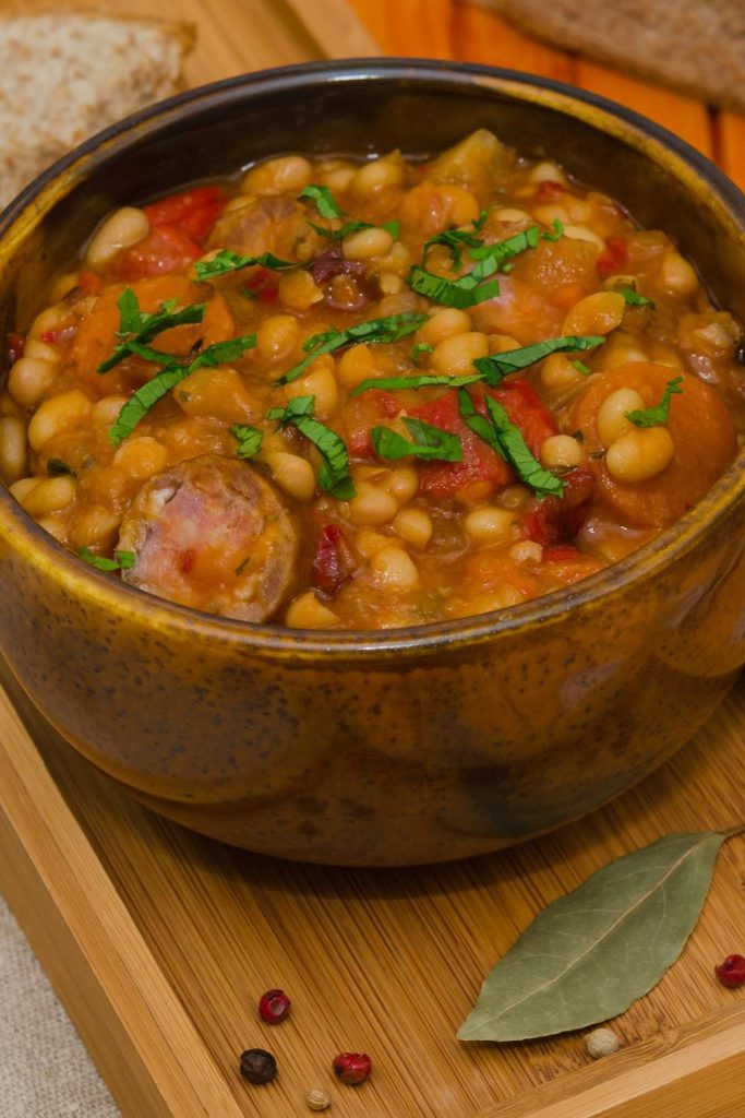 A rustic bowl full of sausage cassoulet, garnished with fresh herbs, alongside a loaf of bread.