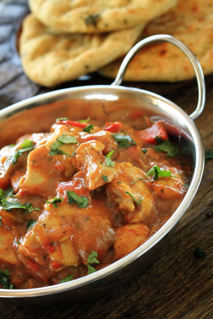 Chicken balti in a metal bowl with naan bread in the background.

