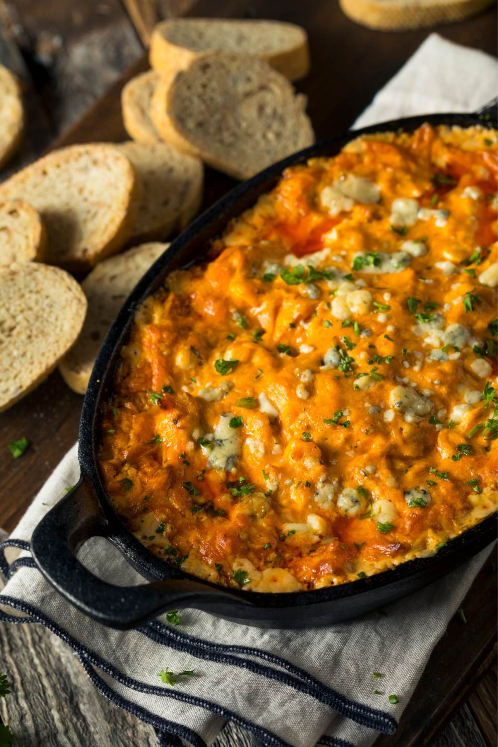 Buffalo chicken dip with a topping of blue cheese and parsley, next to bread slices.