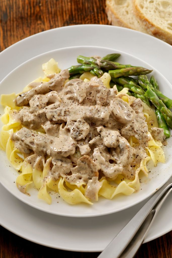 Egg noodles and beef stroganoff with green asparagus on a white plate.