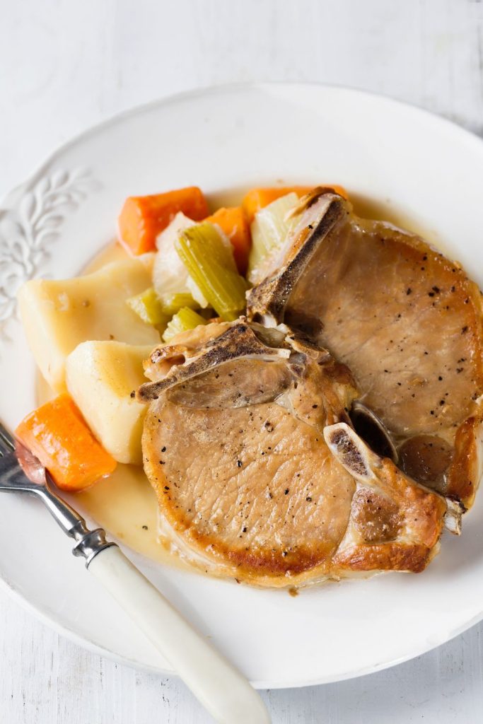 Tender slow cooker pork chops with carrots, celery, and potatoes on a white vintage plate.