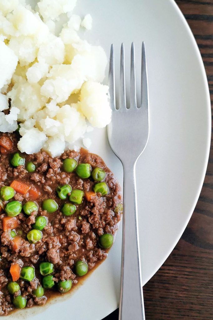 Plate with mince meat, peas, carrots, and mashed potatoes next to a fork.