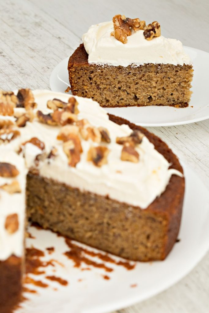 A freshly baked slice of banana bread with cream cheese frosting and walnut topping on a white plate, with the remaining loaf in the background.