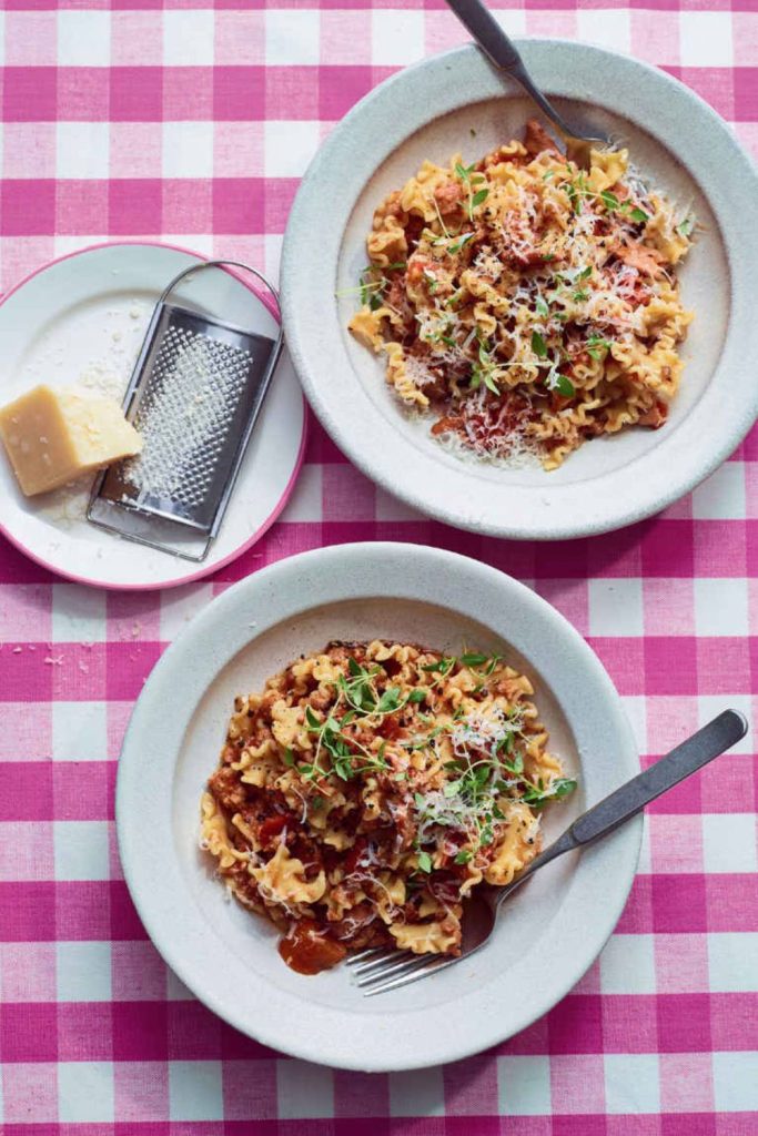Two bowls of slow cooker sausage ragu on a pink checkered tablecloth, cheese grater and parmesan on the side.
