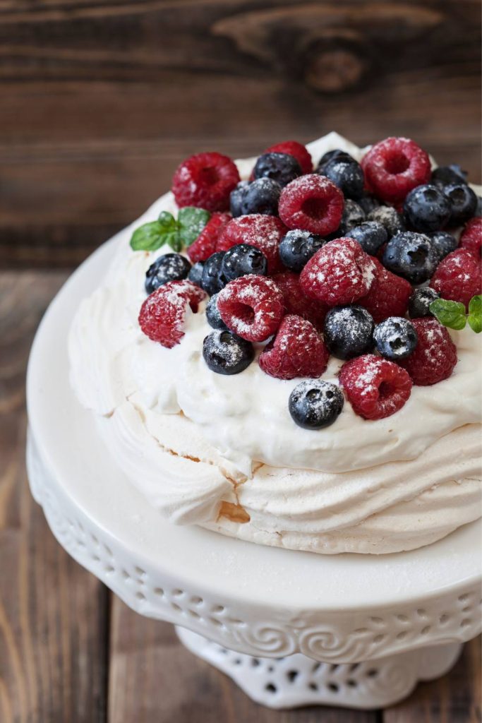 Pavlova topped with whipped cream, raspberries, and blueberries on a cake stand.