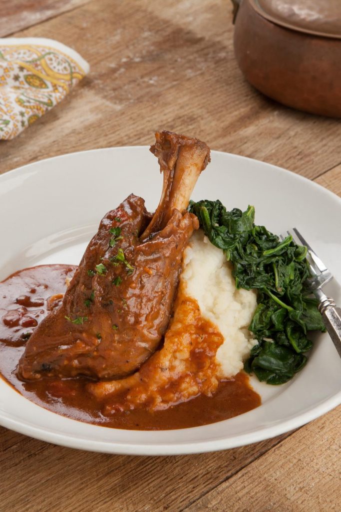 A lamb shank with onion gravy, mashed potatoes, and spinach on a plate.