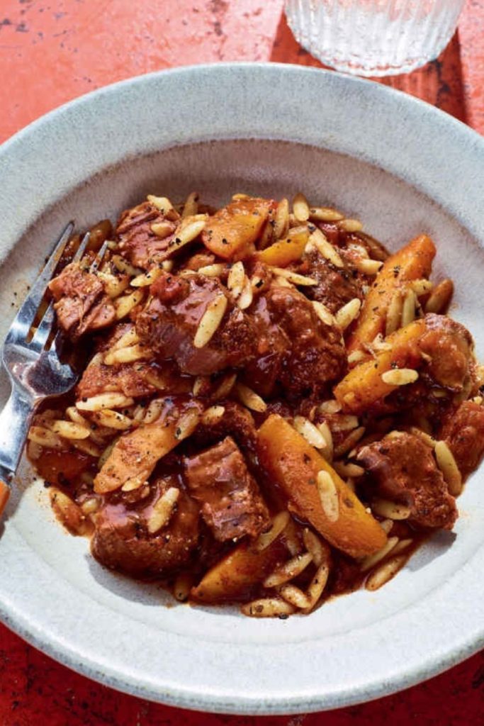 Beef Stifado with orzo and slivered almonds in a grey bowl on a red background.