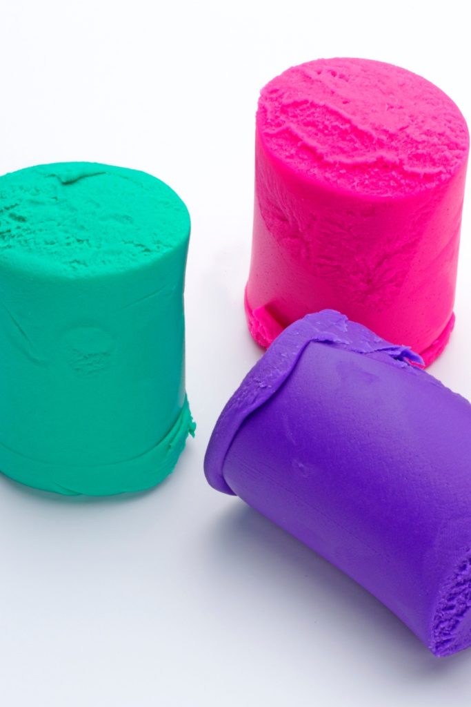 Three colorful playdough cylinders in green, pink, and purple on a white background.