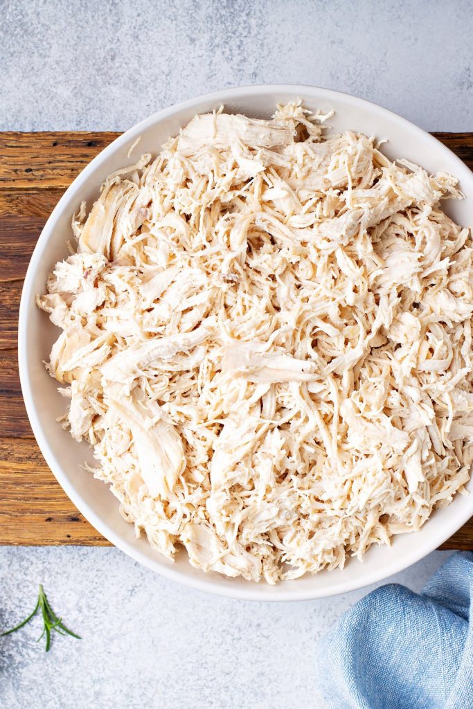 Shredded poached chicken in a white bowl on a wooden board.