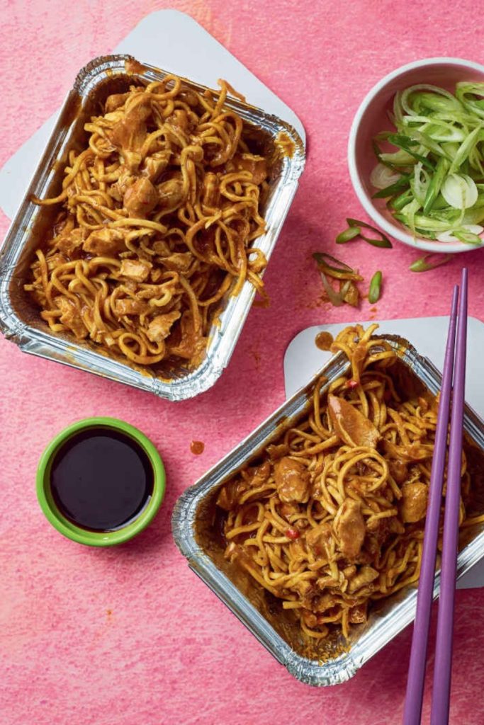 Slow Cooker Chicken Curry Noodles in aluminum containers with chopsticks and green onions on a pink background.