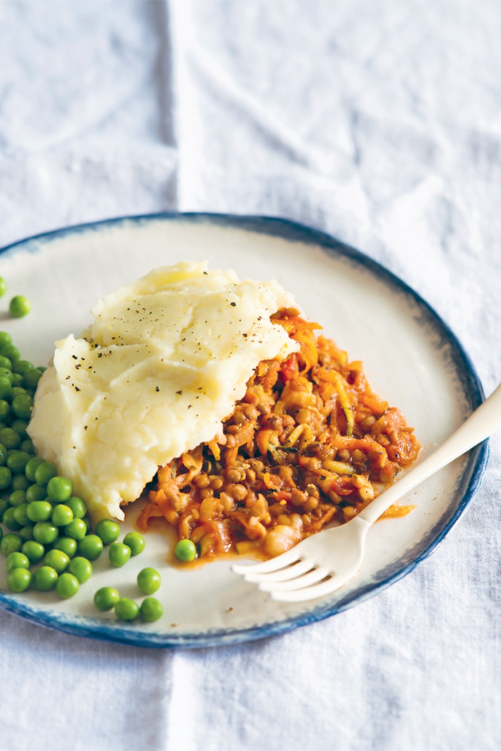 Slow Cooker Lentil Cottage Pie on a plate with mashed potatoes and green peas, served with a fork.