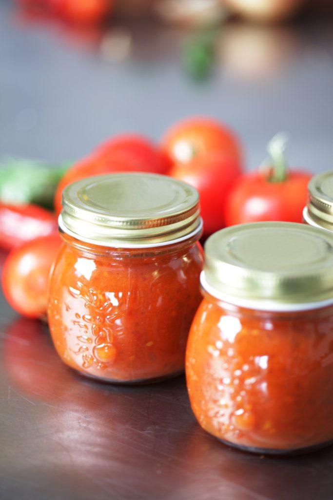 two jars of Slow Cooker Tomato Relish on a table with fresh tomatoes in the background.