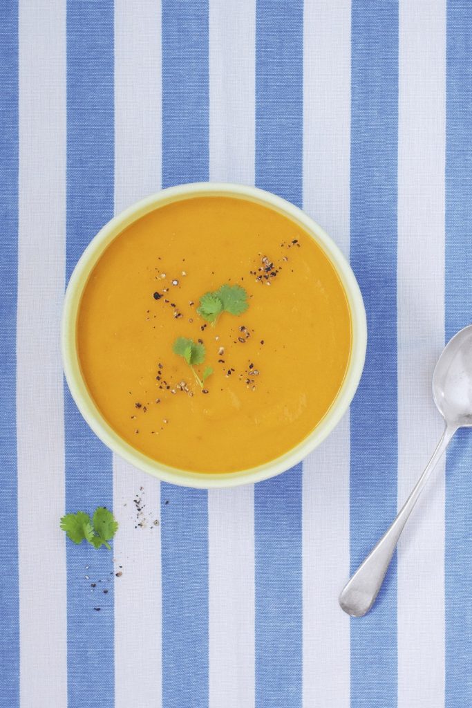 Bright carrot and coriander soup in a yellow bowl on a striped blue and white cloth.