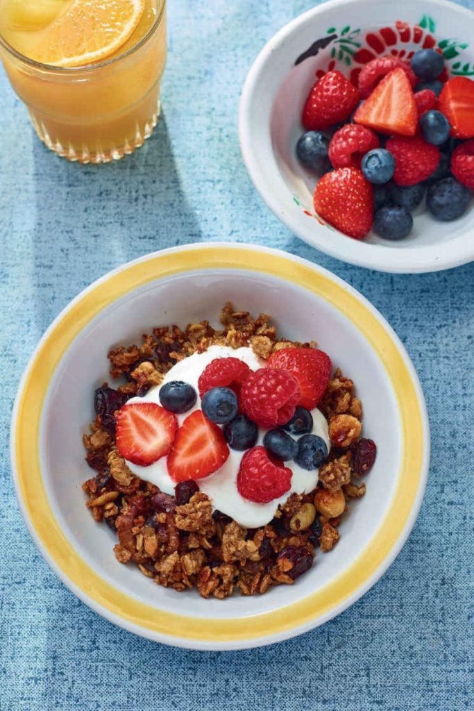 Slow Cooker Granola with yogurt and berries in a yellow-rimmed bowl, alongside a bowl of fresh berries and a glass of orange juice.