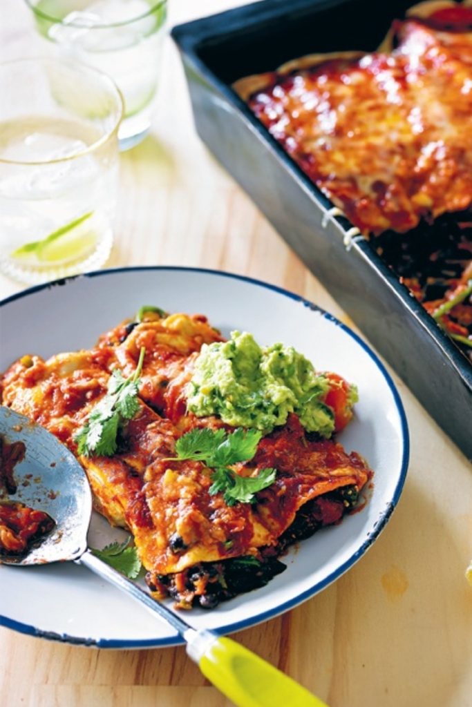 Slow Cooker Black Bean Enchiladas served with guacamole and cilantro on a white plate next to a baking dish.