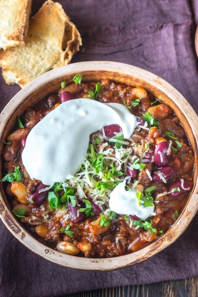 Slow Cooker Elk Chili topped with sour cream, cheese, and herbs in a wooden bowl.