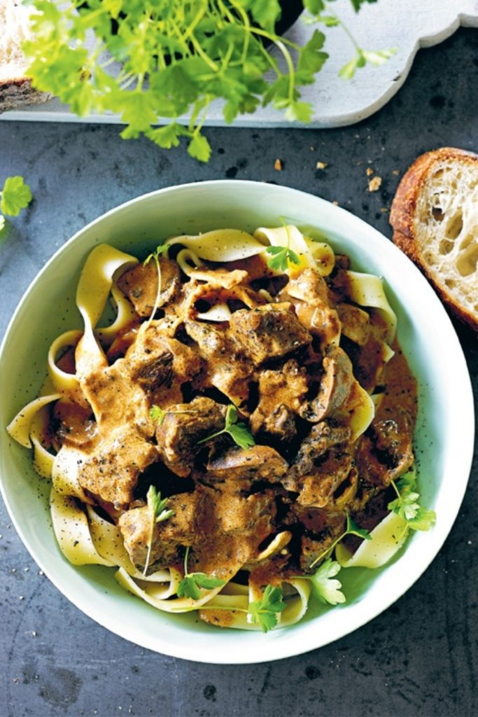 Veal Stroganoff served in a green bowl over pasta, garnished with fresh herbs.