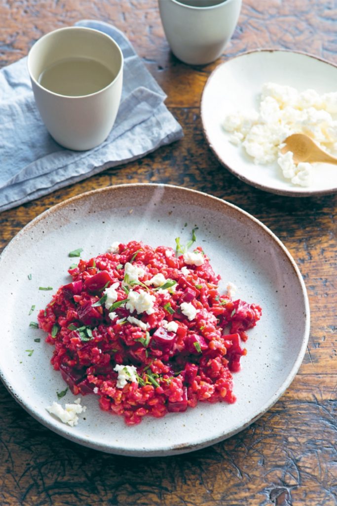Slow Cooker Beetroot Risotto on a plate, garnished with crumbled cheese and herbs, with a cup of drink in the background.