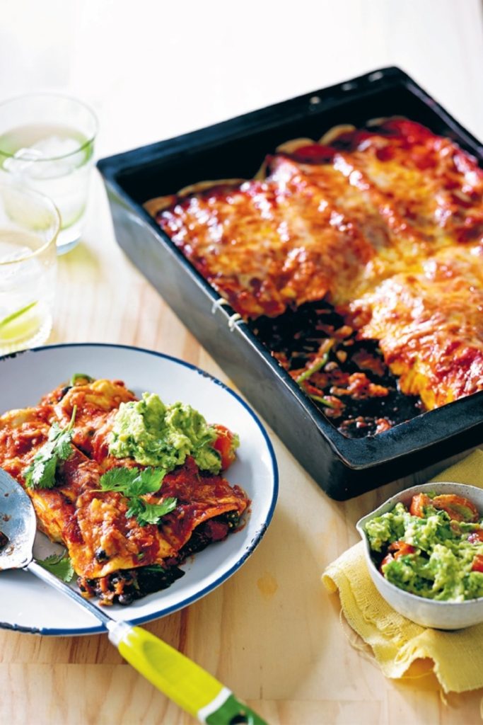 Slow Cooker Black Bean Enchiladas topped with guacamole and cilantro on a white plate with a baking dish in the background.
