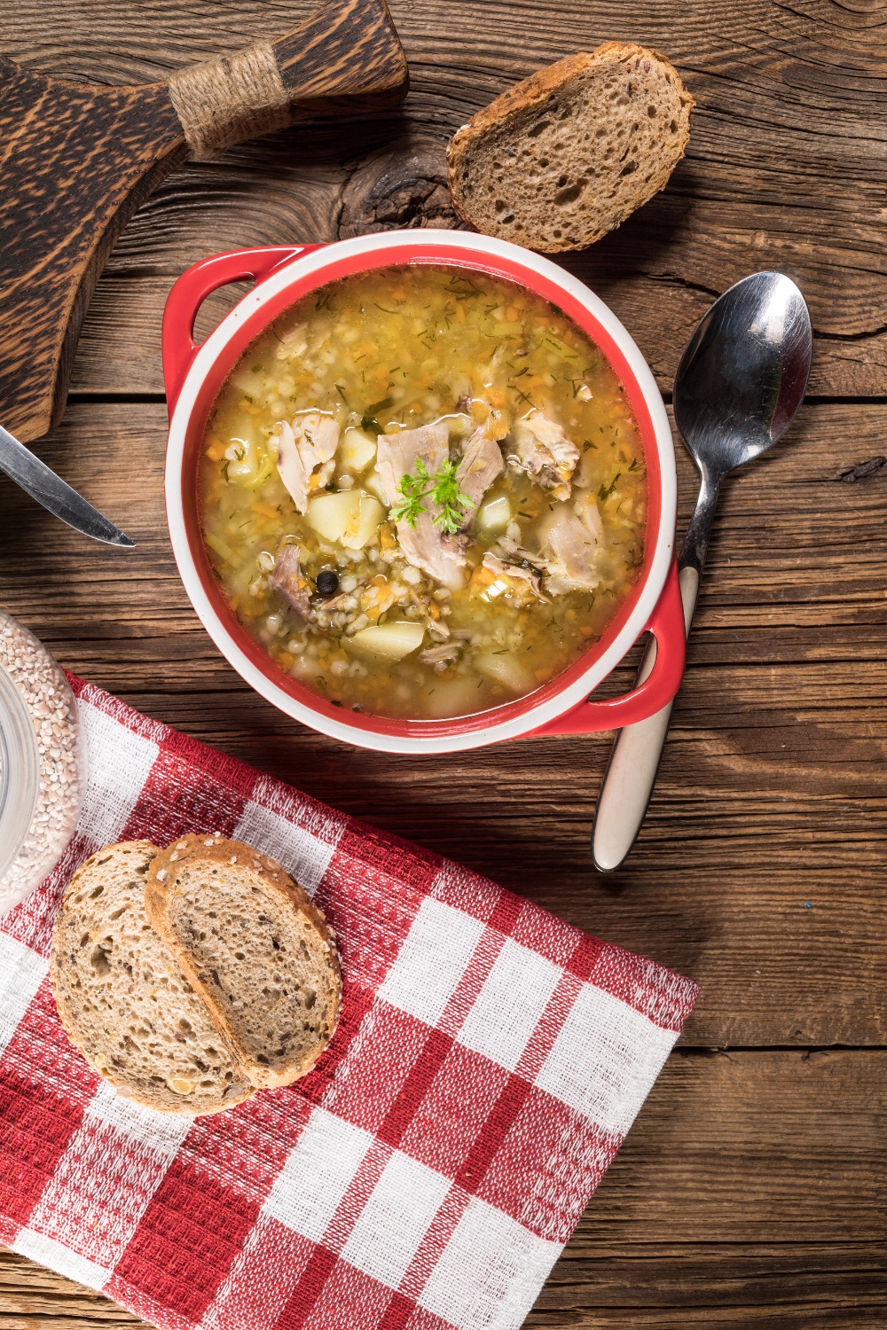 chicken barley soup in a red bowl with a slice of bread on the side.