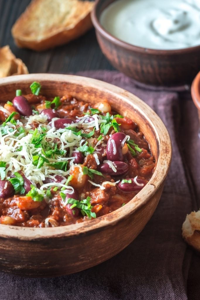 Slow Cooker Elk Chili in a wooden bowl topped with cheese and herbs, with bread and sour cream on the side.