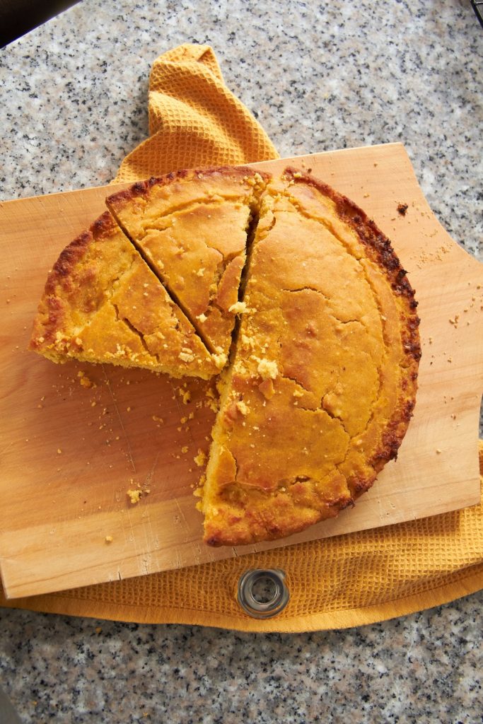 A round cornbread on a wooden cutting board, partially sliced.