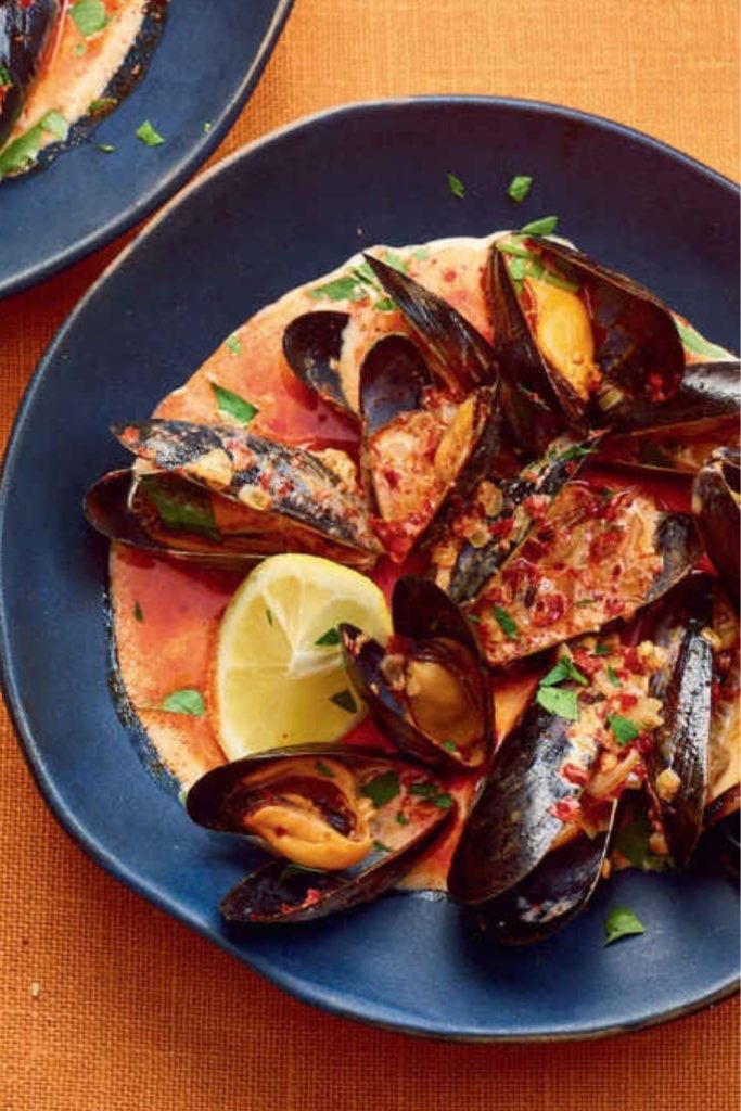 mussels in a tomato-based sauce with lemon wedges on a blue plate.