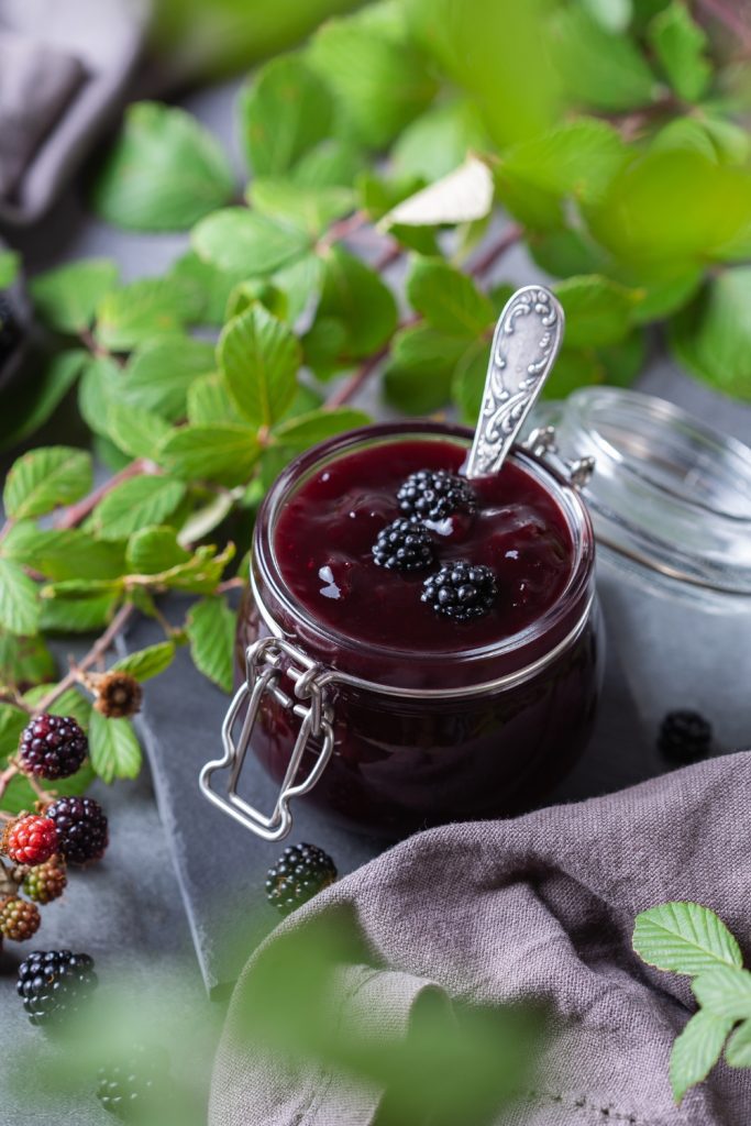  Jar of blackberry jam with fresh blackberries and a wooden bowl in the background.