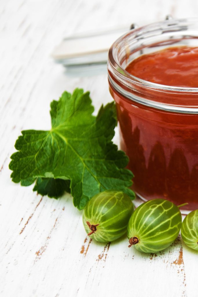 Jar of gooseberry jam with green gooseberries and leaves on a white wooden surface.