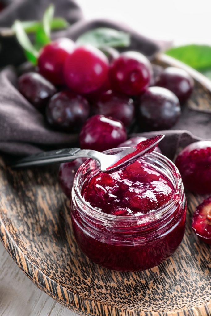 A jar of plum jam with a basket of fresh plums and a red-checkered cloth.