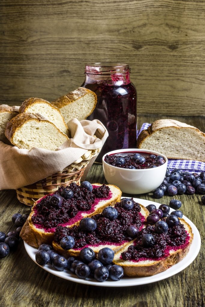 Two small jars of Slow Cooker Blueberry Jam on a wooden surface with fresh blueberries and leaves.