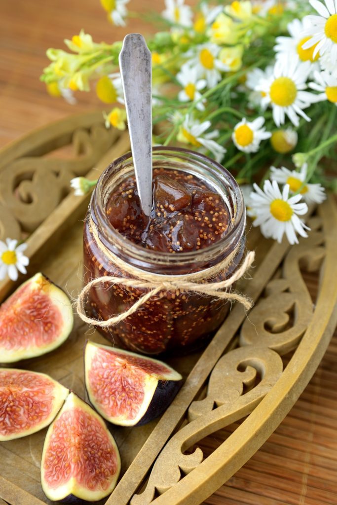 Jar of fig jam with a spoon and fresh figs on a tray with flowers.