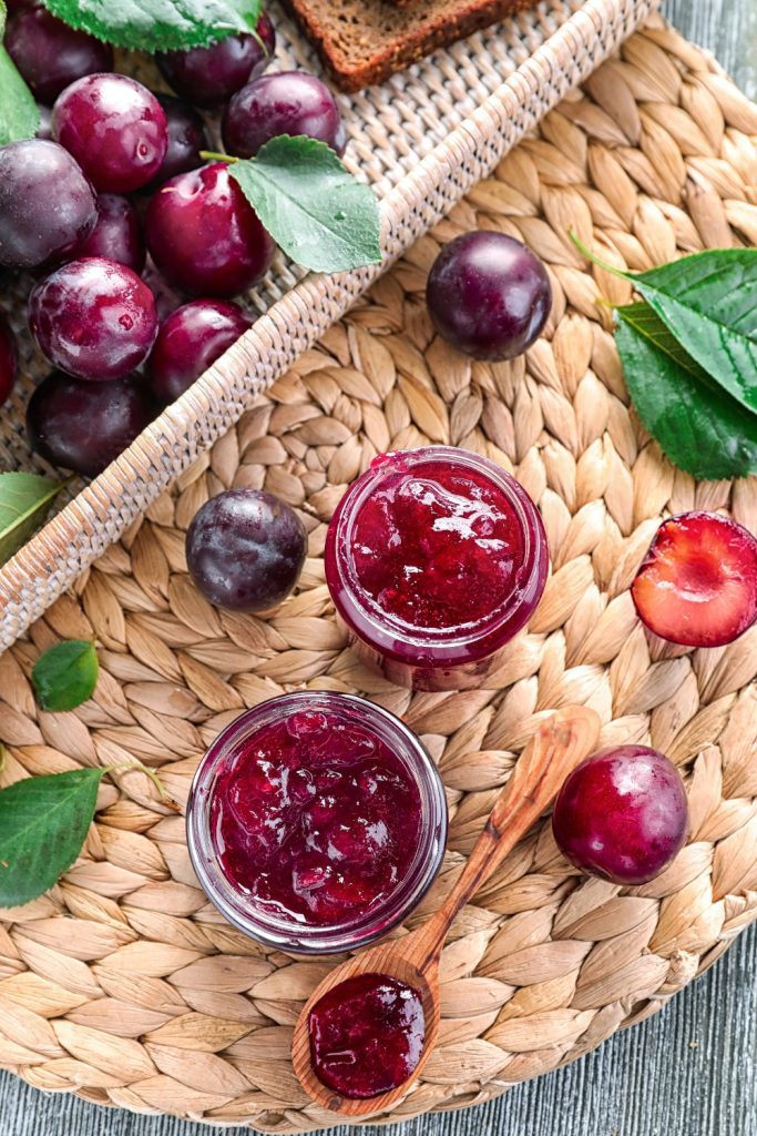 Two jars of plum jam on a woven mat with fresh plums and a wooden spoon.