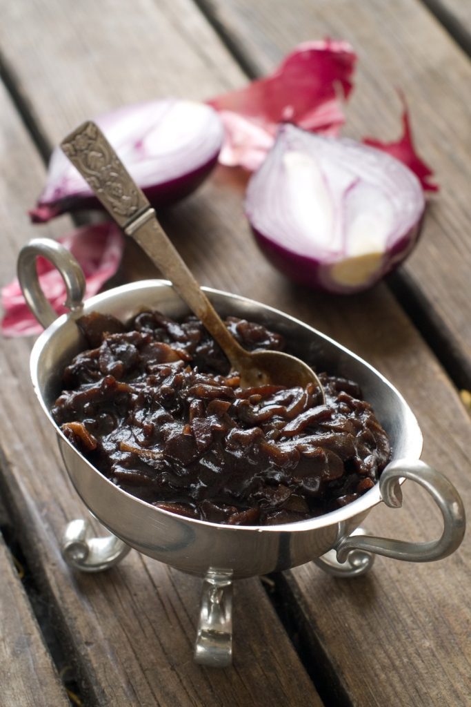 Slow Cooker Onion Jam in a silver serving dish with red onions in the background.