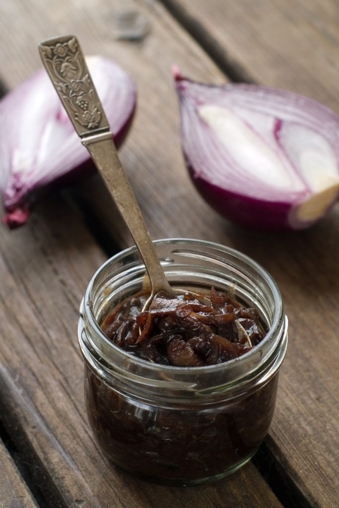 Slow Cooker Onion Jam in a jar with a spoon, with red onions in the background.