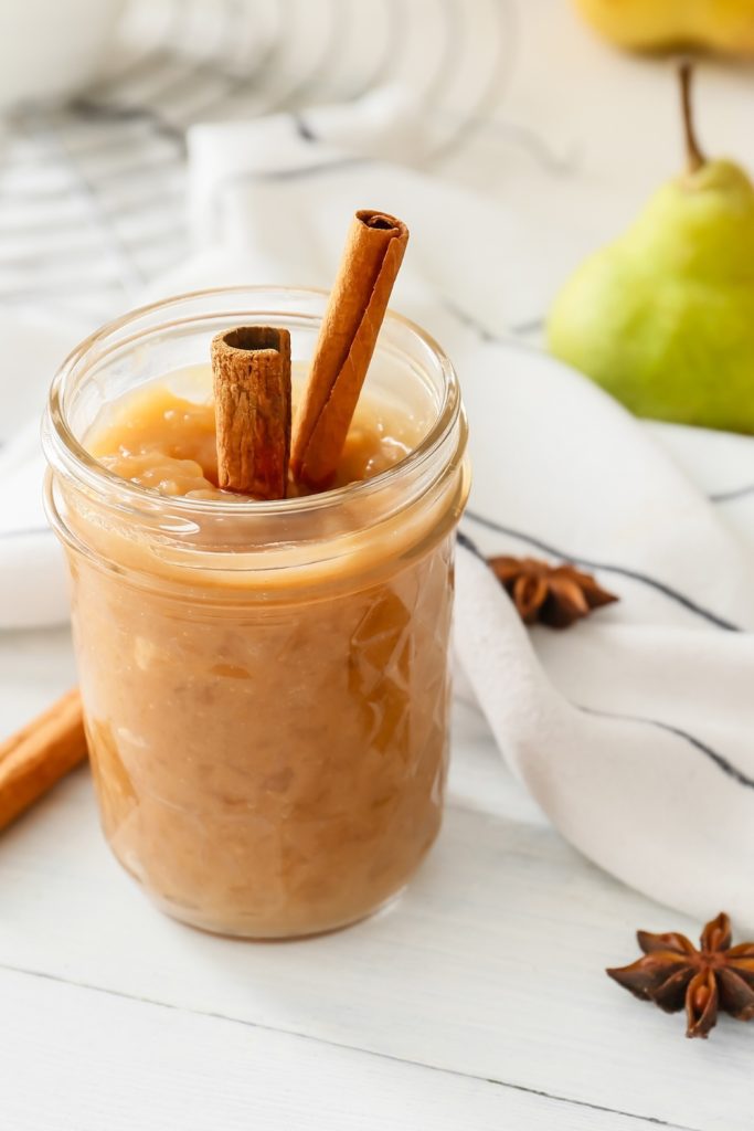 Slow Cooker Pear Jam in a jar with two cinnamon sticks, with a pear in the background.