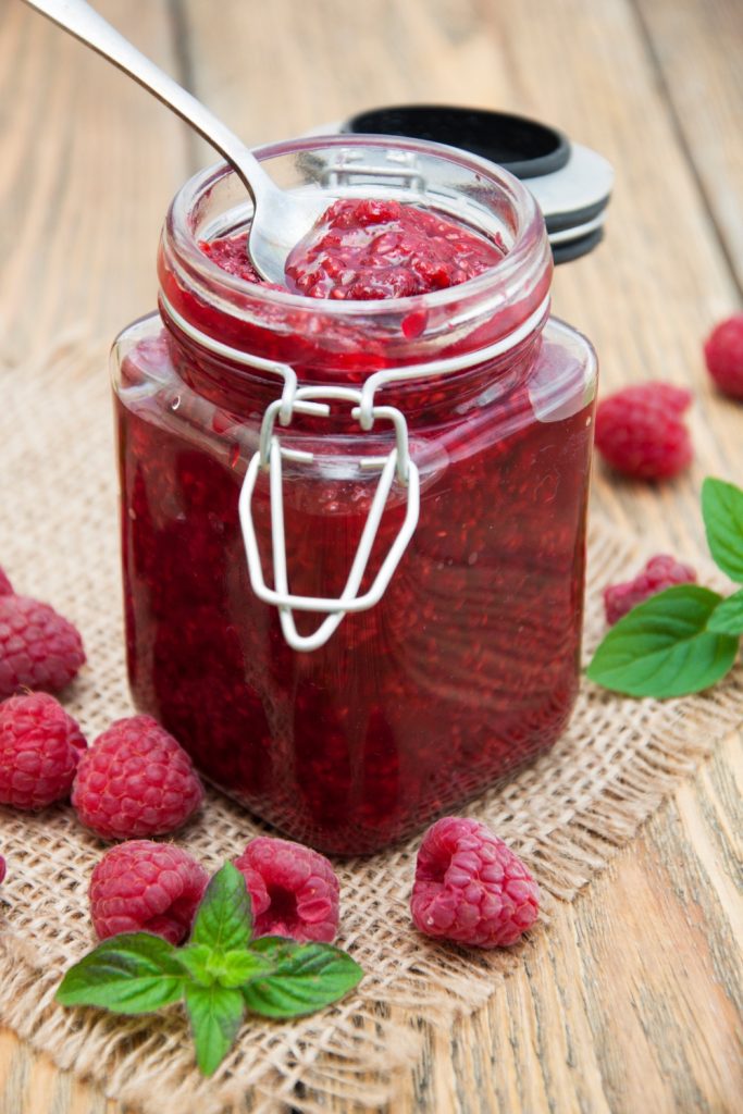 Jar of raspberry jam with fresh raspberries on a wooden surface.