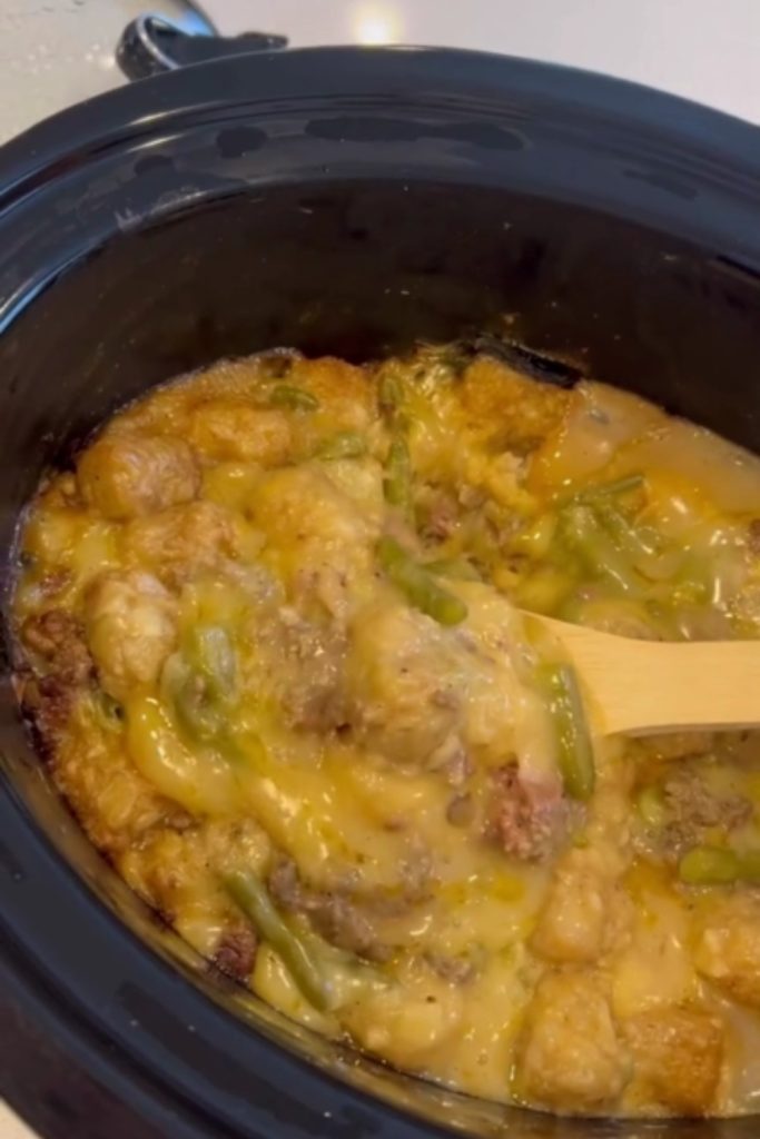 Slow Cooker Tater Tot Casserole being stirred with a wooden spoon, showing melted cheese and green beans.