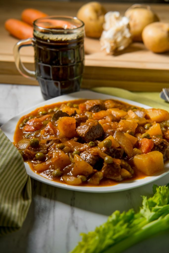 Jumbuck Stew served in a white plate with a glass of dark beer and vegetables in the background.