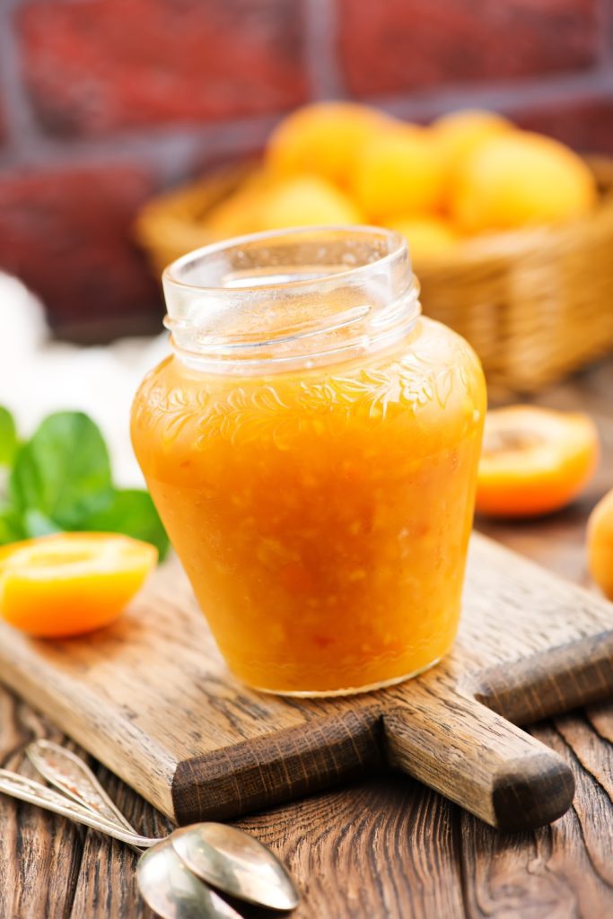 Jar of apricot jam on a wooden board with fresh apricots in the background.