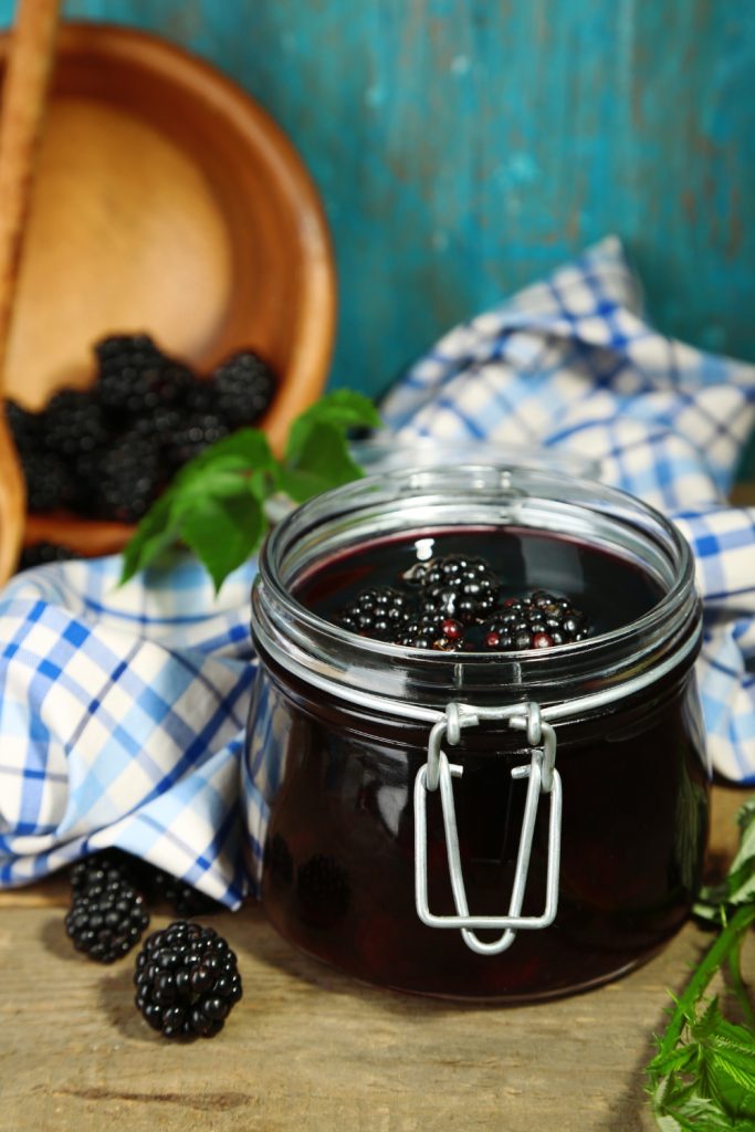 Jar of blackberry jam on a wooden board with fresh blackberries and green leaves.