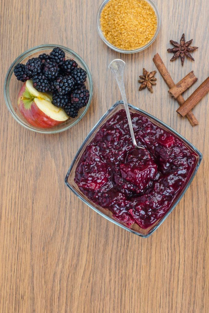 Bowl of blackberry and apple jam with fresh blackberries, apple slices, brown sugar, cinnamon sticks, and star anise on a wooden table.