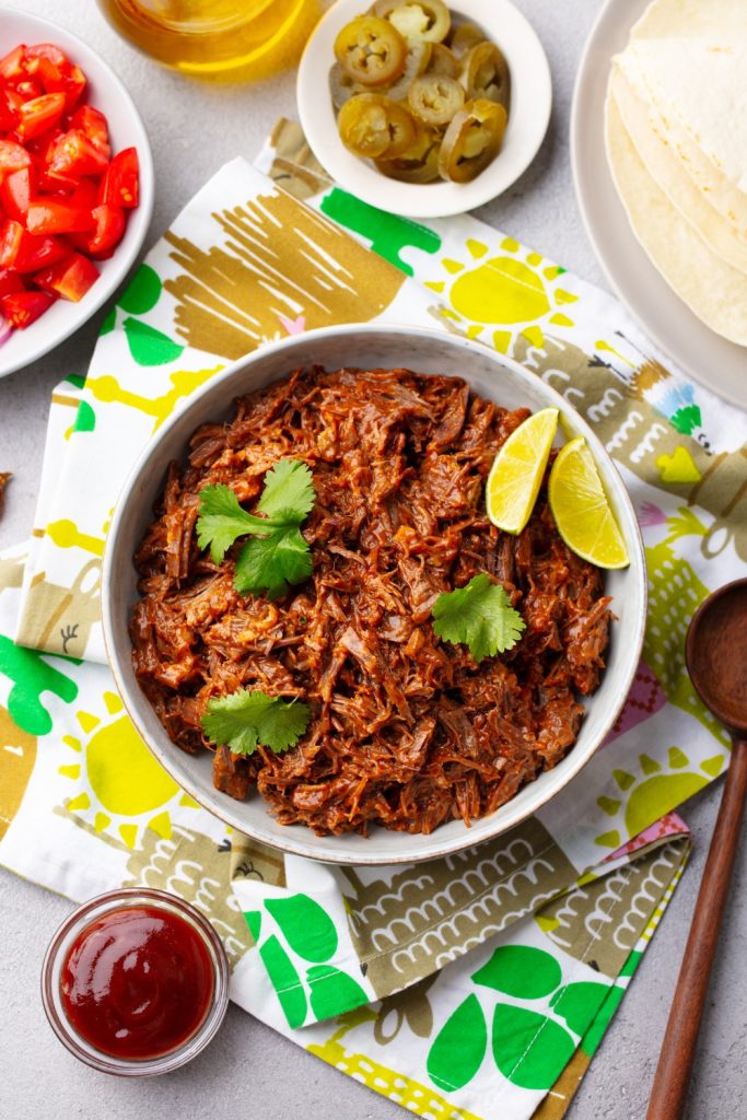 slow cooker carne mechada with garnishes, tortillas, and side dishes.